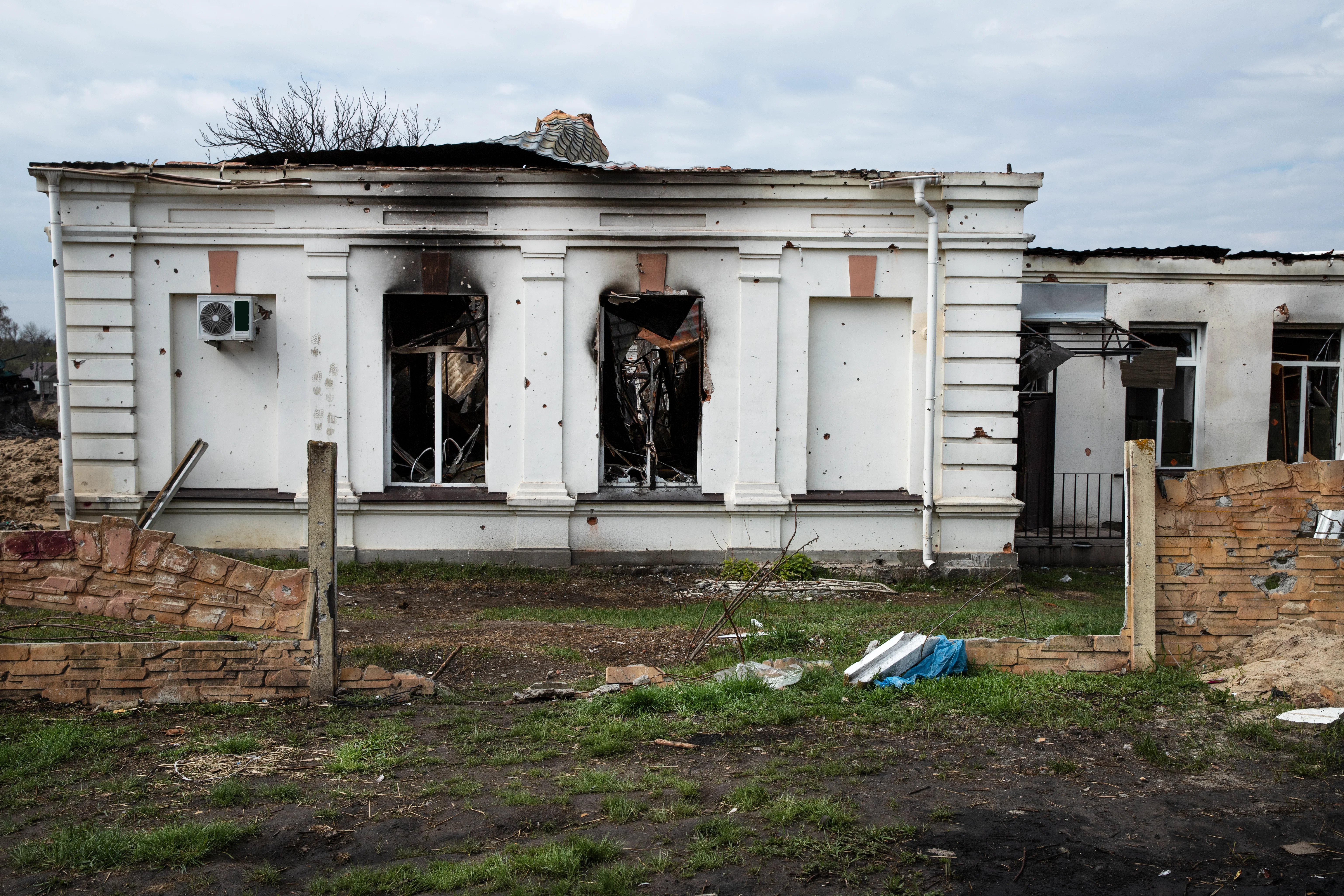 A white building with bullet holes, burned-out window frames, a damaged roof, and a broken brick fence.