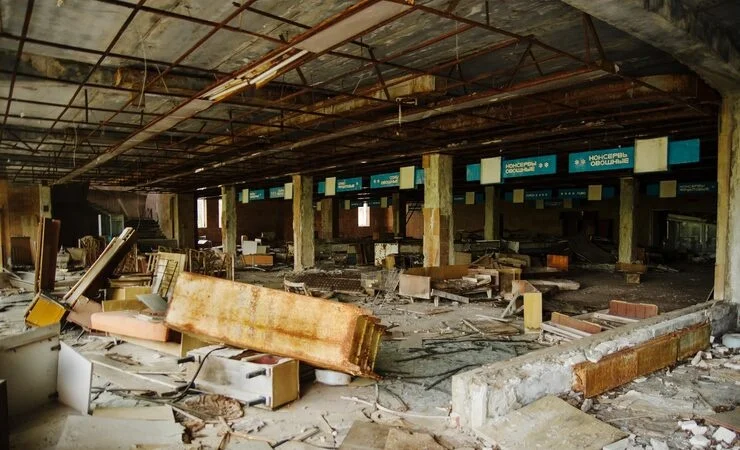 A wide shot of a severely dilapidated and abandoned supermarket interior, with debris scattered across the floor and rusty fixtures on the ceiling.
