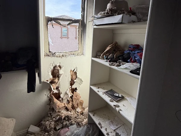 A severely damaged room with exposed wall insulation, a broken window revealing another building, and shelves covered in debris.