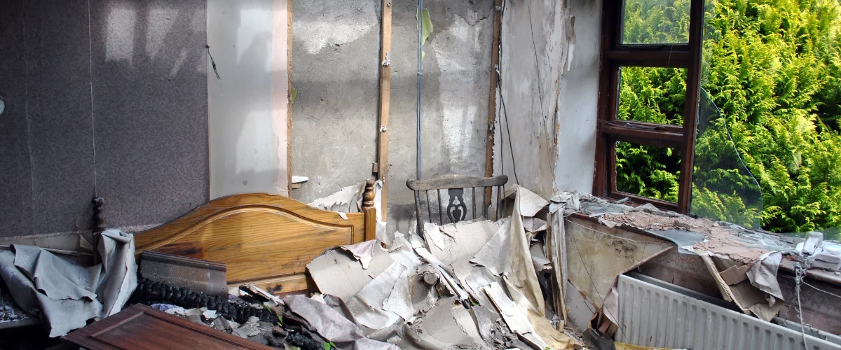 A desolate room after a fire, showing peeling wallpaper, a charred bed, broken window, and scattered debris.