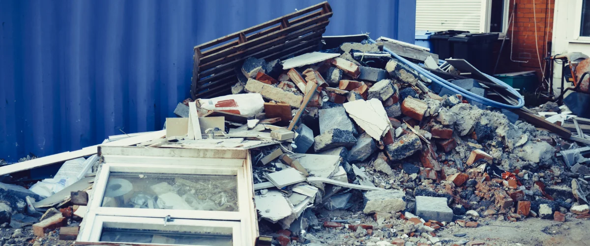 A large pile of construction debris, including bricks, concrete, and a window frame, sits against a blue container. A large pile of construction debris, including bricks, concrete, and a window frame, sits against a blue container.