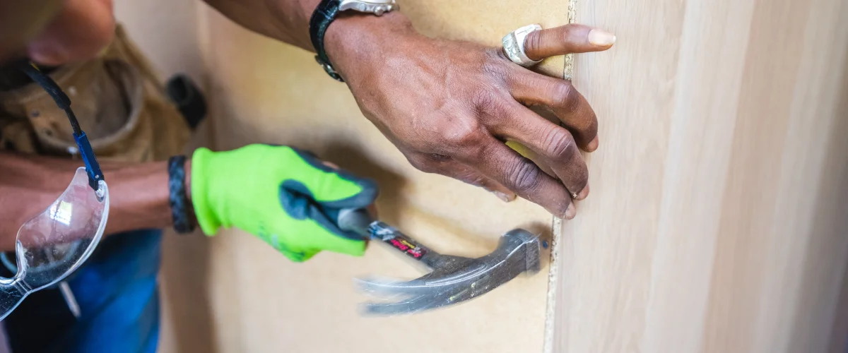 A craftsman in bright green gloves hammers a nail into light wood, with safety glasses hanging nearby. A craftsman in bright green gloves hammers a nail into light wood, with safety glasses hanging nearby.