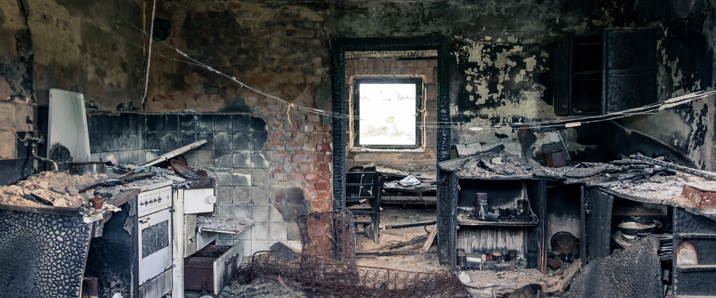 A wide view of a severely fire-damaged, burnt-out kitchen with debris, broken appliances, and charred walls.