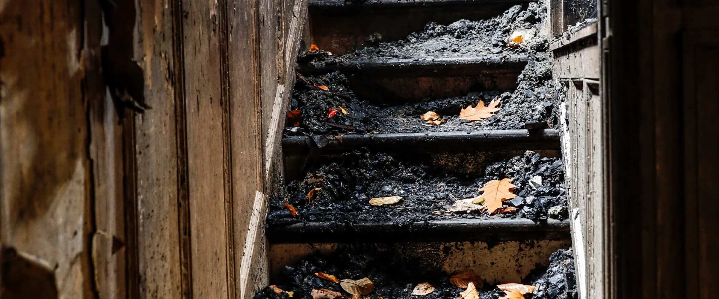 A dark, damaged wooden staircase covered in black char and debris, with scattered autumn leaves on the steps.