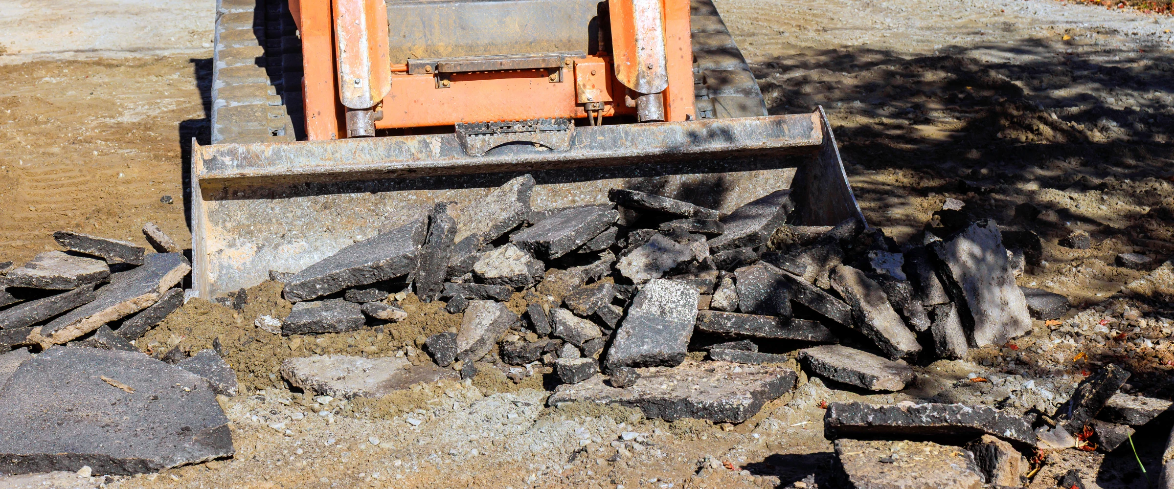 An orange excavator bucket filled with broken pieces of black asphalt on a dirt ground at a construction site. An orange excavator bucket filled with broken pieces of black asphalt on a dirt ground at a construction site.