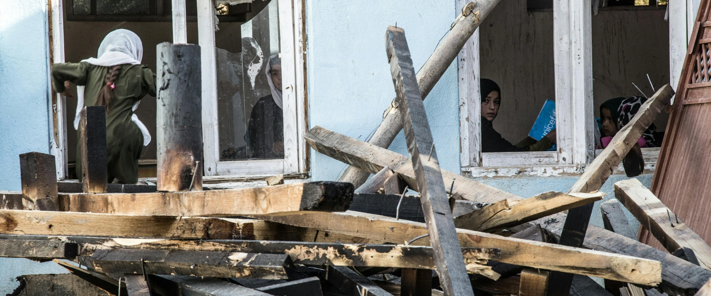 Children, some wearing hijabs, look out from broken windows of a blue building, with debris and wooden planks in the foreground. Children, some wearing hijabs, look out from broken windows of a blue building, with debris and wooden planks in the foreground.