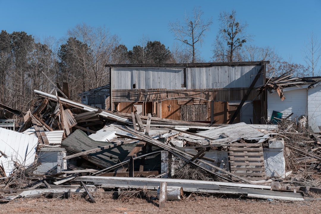 A severely damaged brick house with its roof destroyed and debris scattered, against a bright blue sky with clouds.