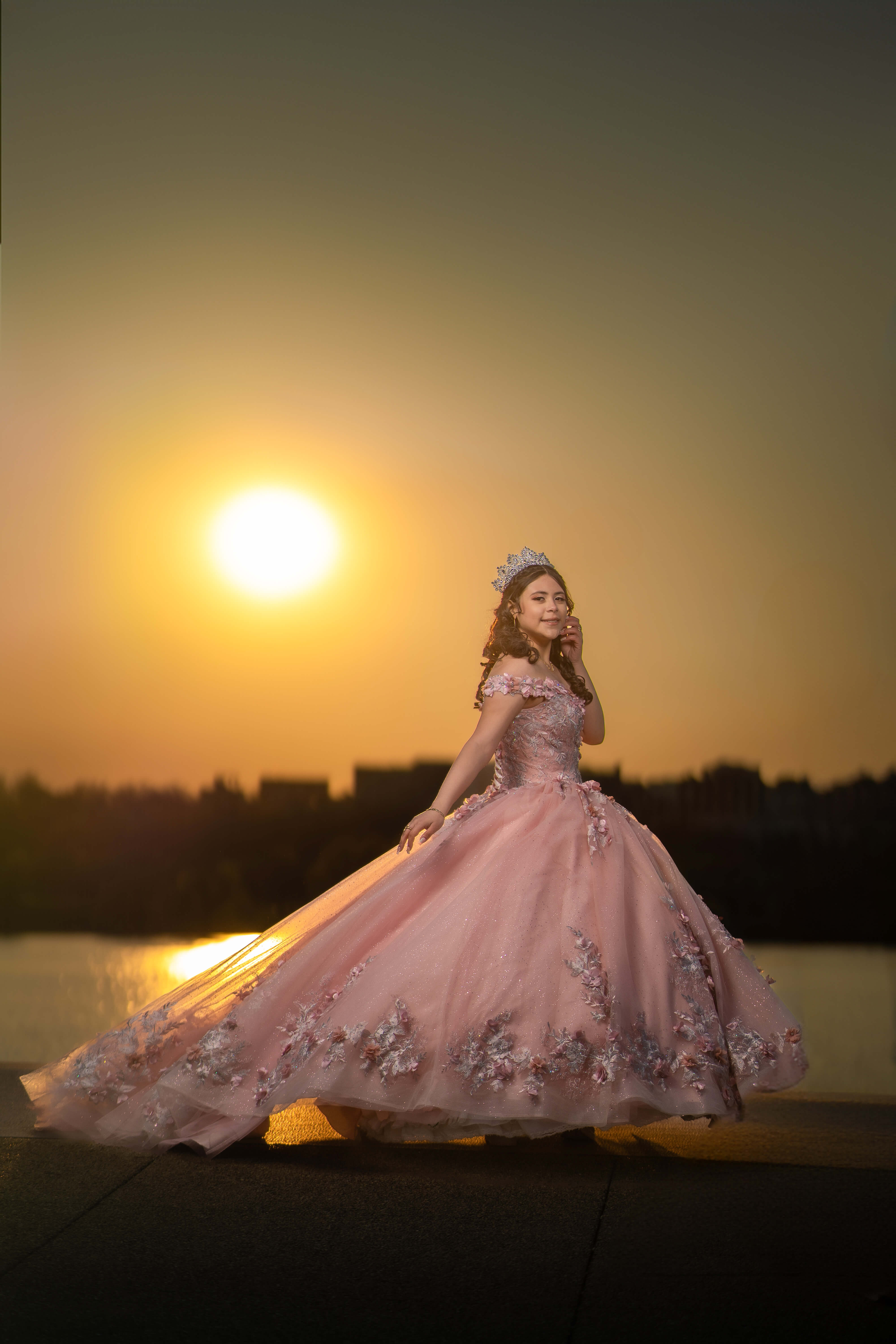 Quinceañera con vestido rosa y corona al atardecer junto al agua