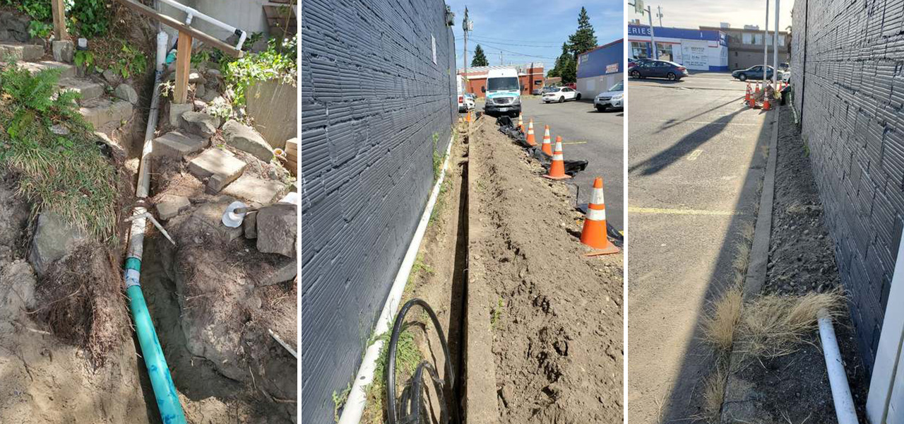Outdoor drainage pipes and trench work along a black brick wall, safety cones.