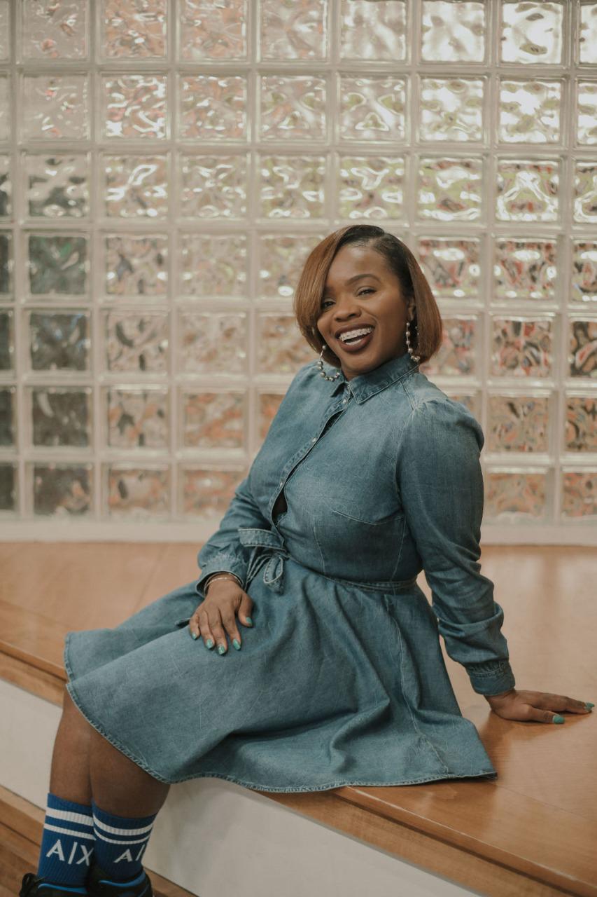 Koach Keisha sitting on a wooden step, smiling confidently. She is wearing a stylish denim dress with a cinched waist, accessorized with hoop earrings and bracelets. Her hair is neatly styled in a sleek bob, and she has braces on her bright smile. Her nails are painted a vibrant blue, matching her blue AIX socks. The background features a wall of textured glass blocks, creating a modern and artistic setting.
