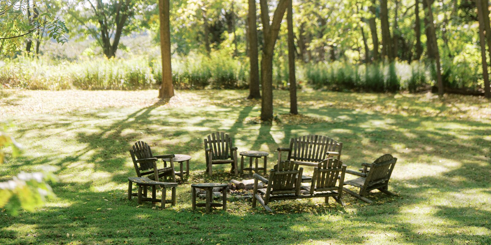 A circle of weathered wooden chairs set in dappled light on a lawn by the water.