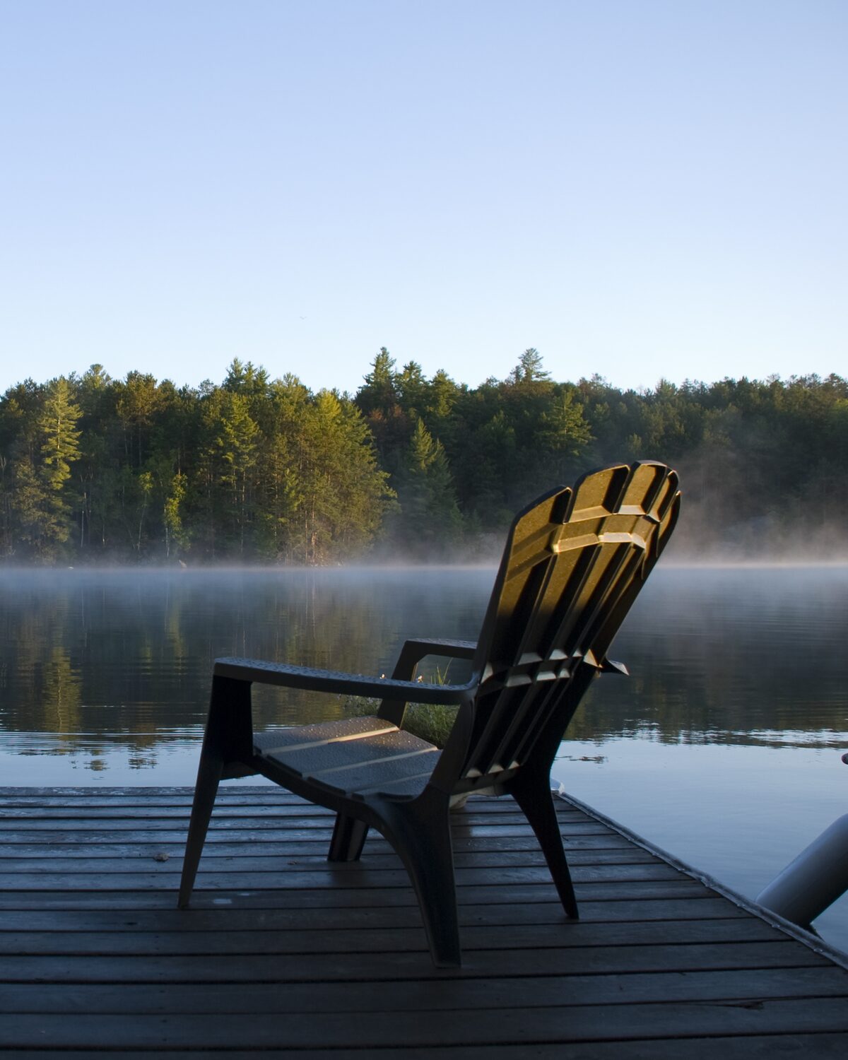 An empty wooden chair at the end of a dock, facing a misty lake in morning light.