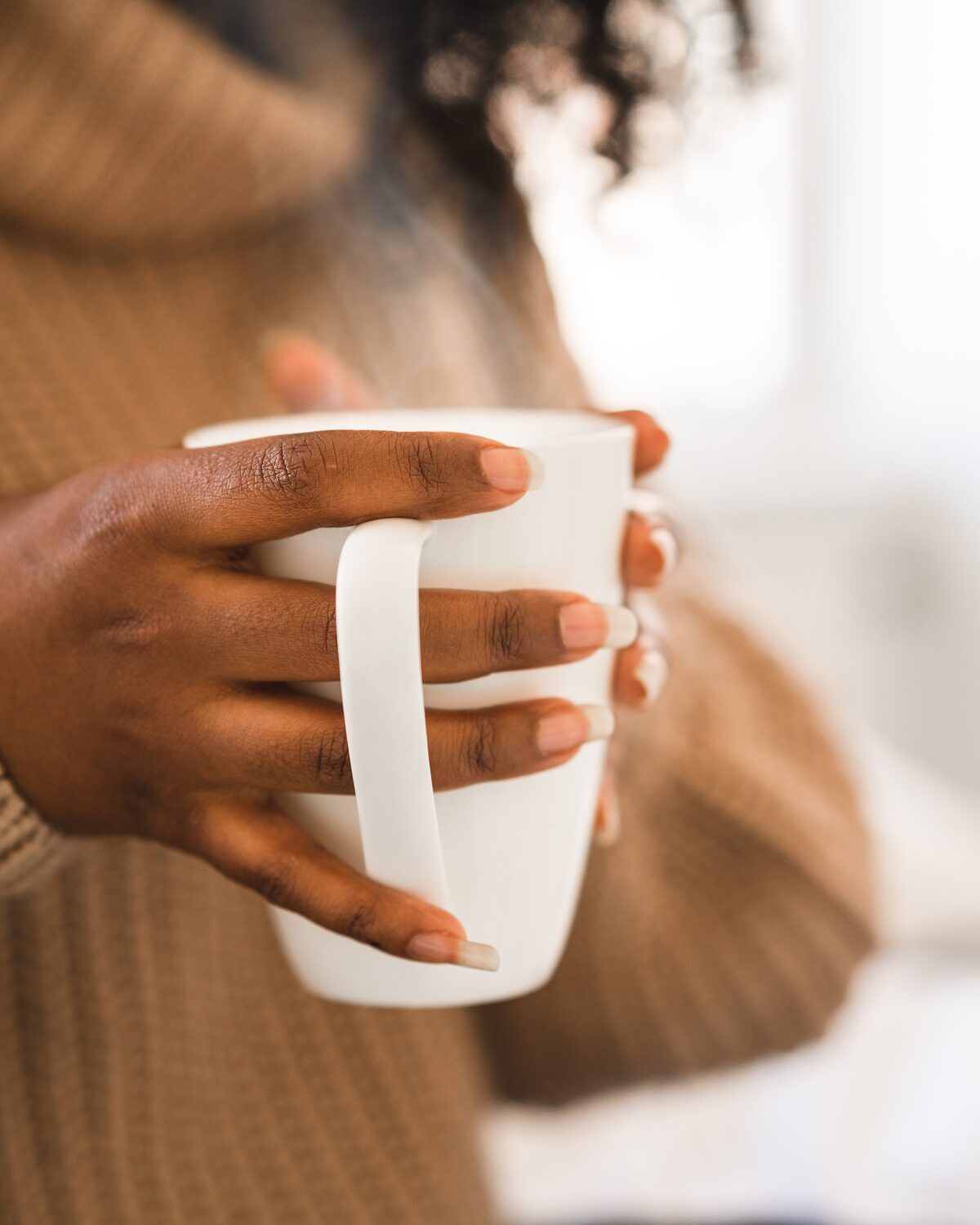 A woman in a soft sweater holding a white mug in both hands, morning light on her face.