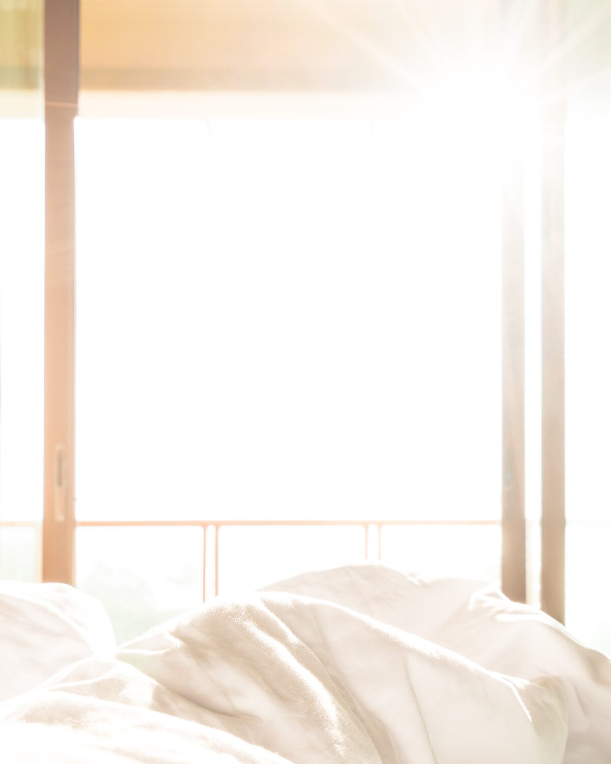 A bed with white linens below a tall window, morning light coming through the curtains.