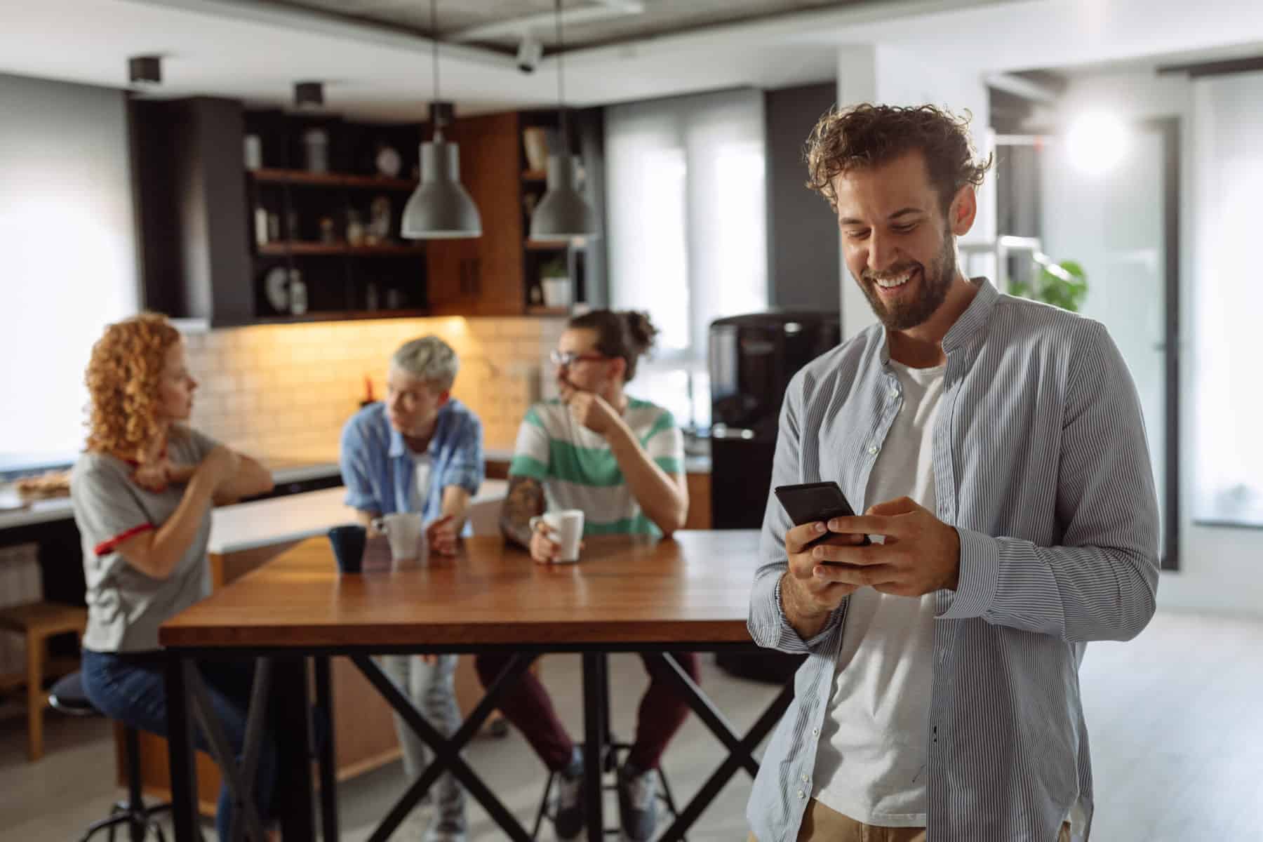 Man texting in kitchen with friends nearby.