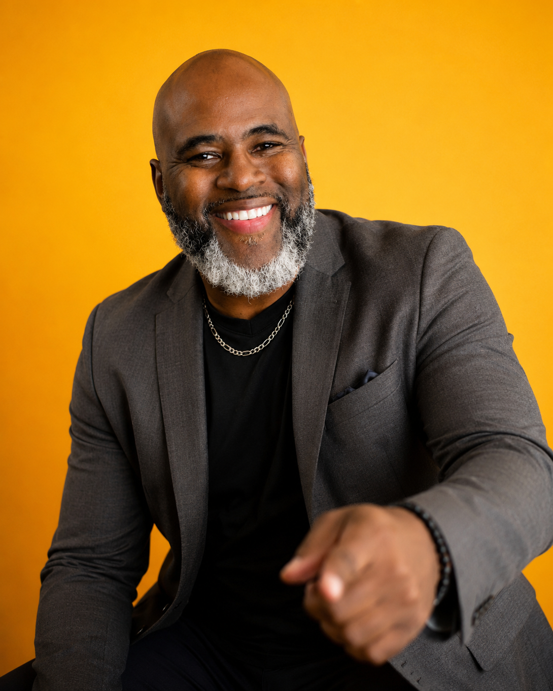 Kelly James smiling and pointing toward the camera, wearing a charcoal jacket against a golden yellow background