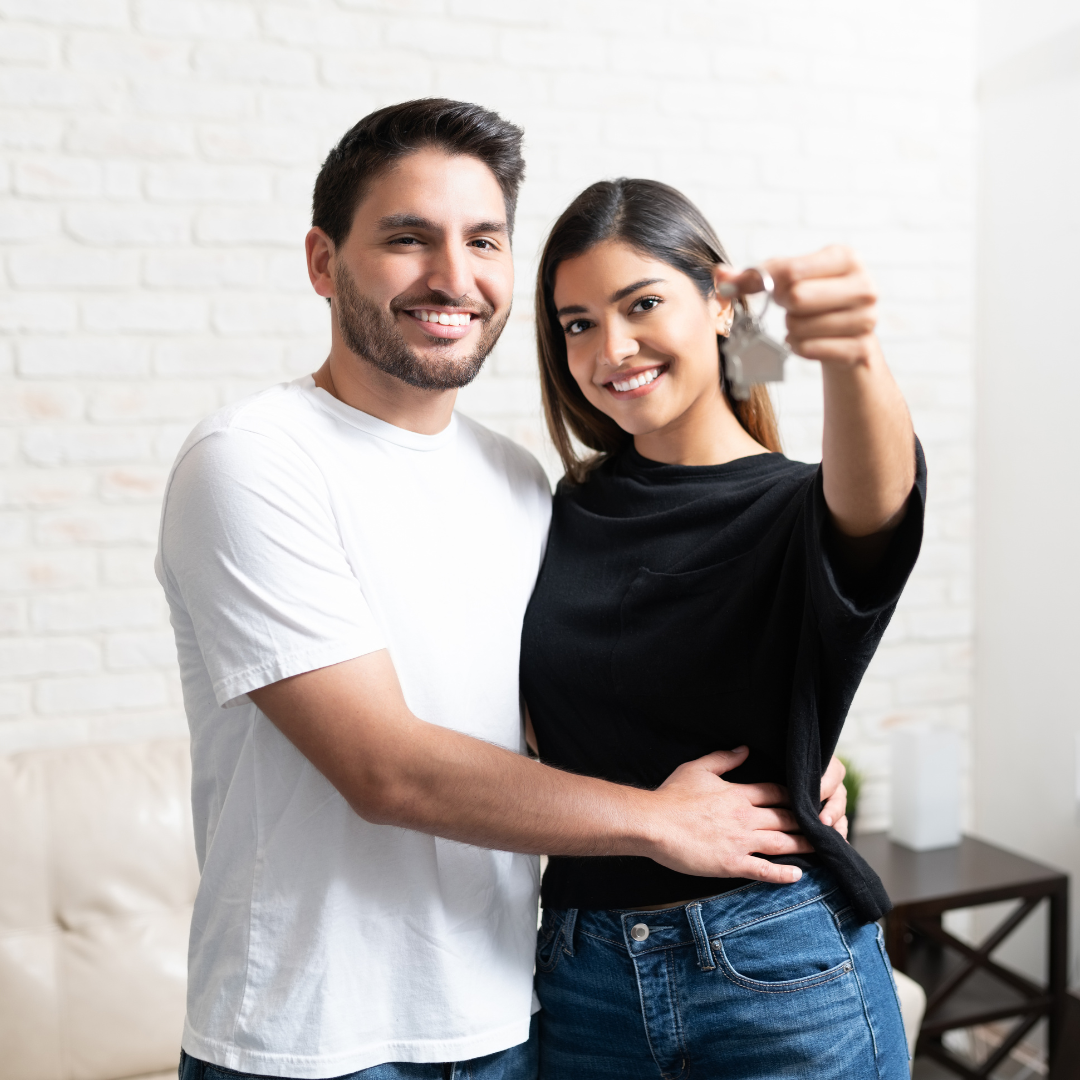 Pareja latina joven frente a su primera casa en Ontario, sosteniendo unas llaves, con expresión natural de alegría