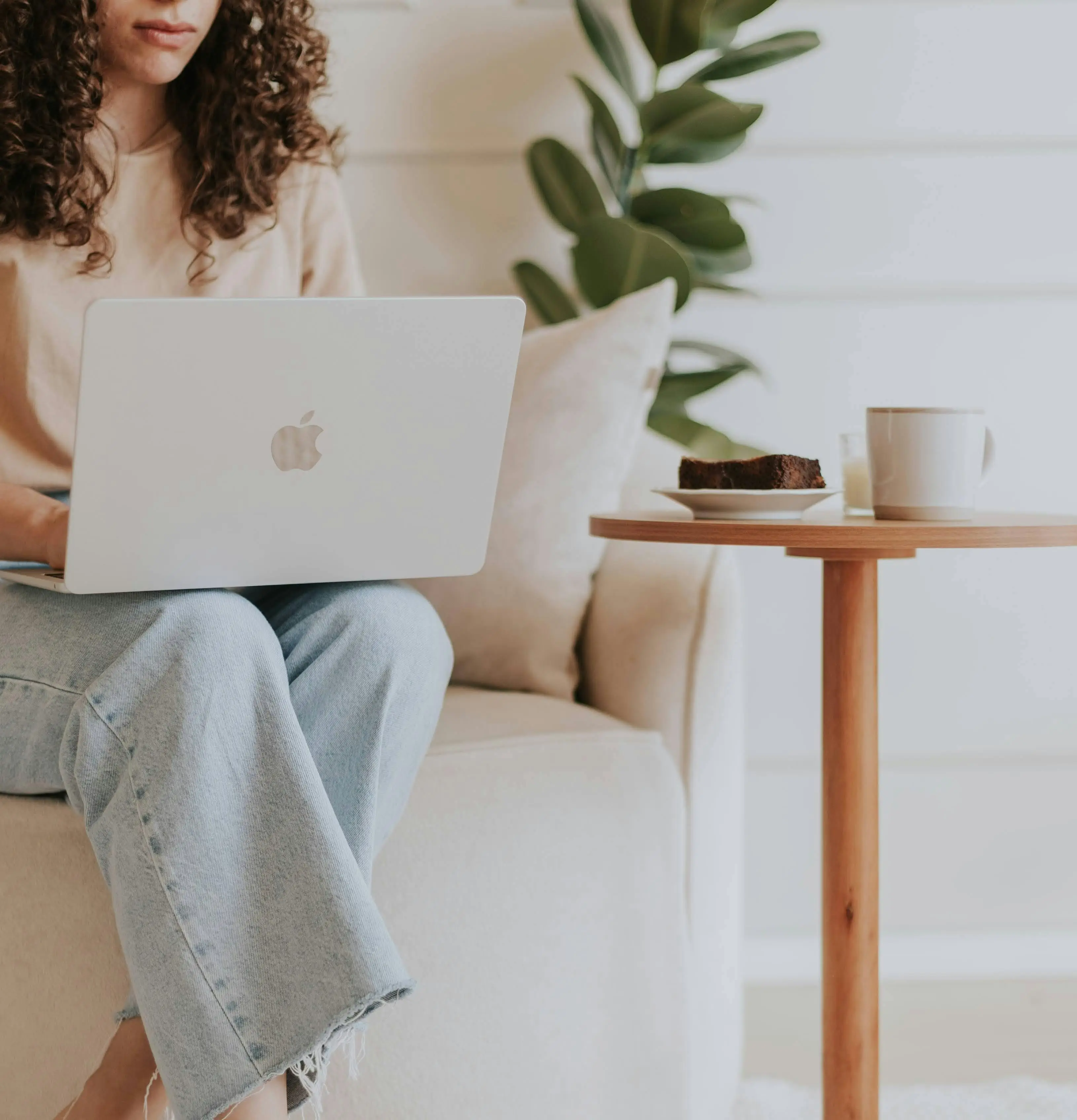 woman sitting on a couch in a neutral room with warm light working on her laptop with a plant and coffee cup nearby