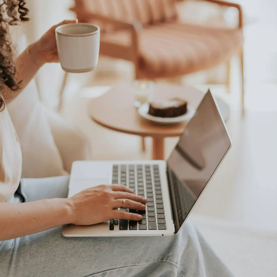 Woman sitting on a couch with a coffee cup in one hand and using her laptop on the other