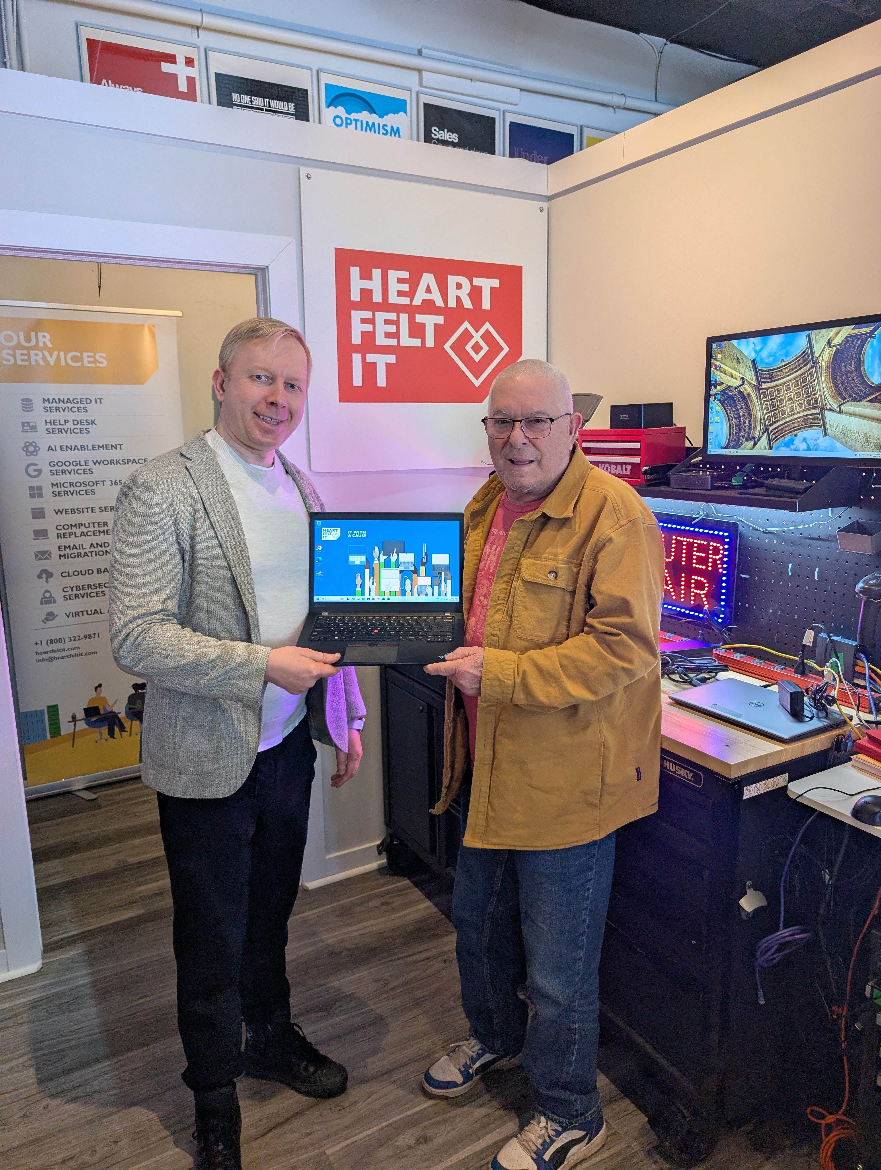 **Alt Text (SEO Optimized):** Heartfelt IT Laptop Giveaway in Kanata: An elderly recipient smiles while receiving a free refurbished business laptop from Heartfelt IT founder Sergey. They are in the IT workshop with the red geometric heart logo visible. The donation bridges the digital divide for local seniors and nonprofits.