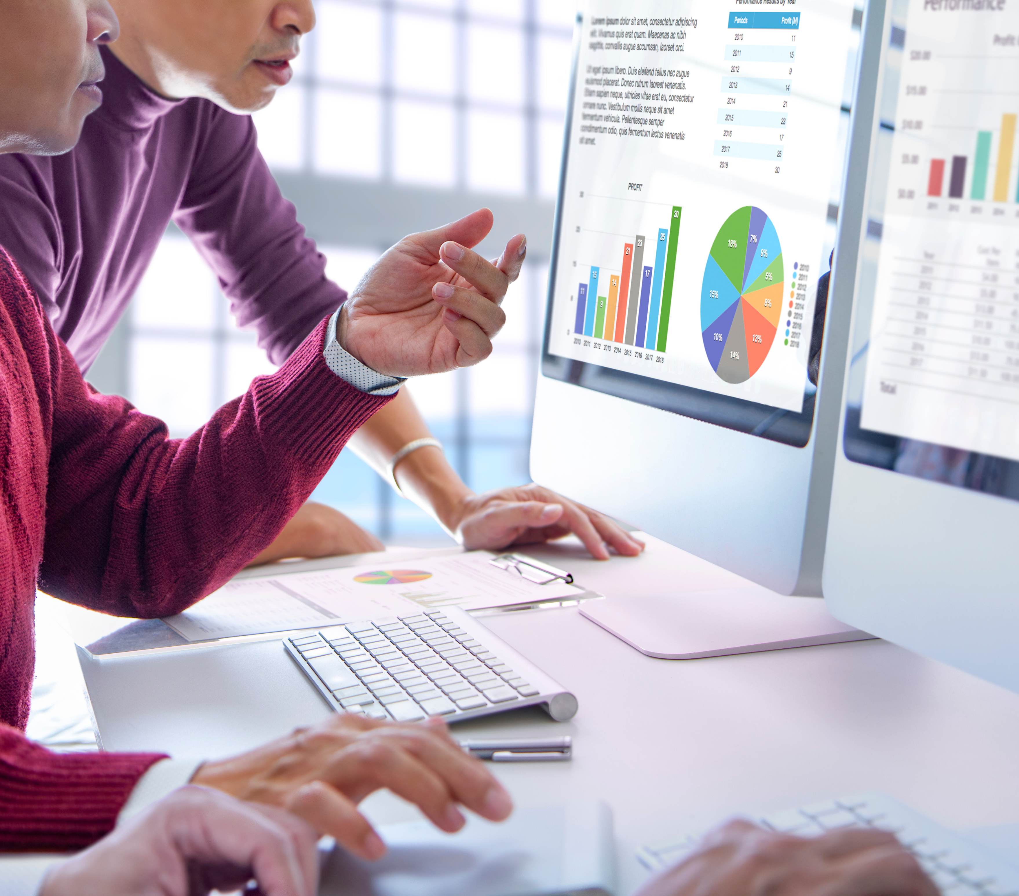 An office scene showing two people looking at a computer screen that displays data charts and graphs. One person, wearing a maroon sweater, points at the screen while the other person, in a purple turtleneck, looks on. On the desk, there is a keyboard, a mouse, a pen, and a clipboard with printed reports. Another monitor is partially visible on the right side of the frame. An office scene showing two people looking at a computer screen that displays data charts and graphs. One person, wearing a maroon sweater, points at the screen while the other person, in a purple turtleneck, looks on. On the desk, there is a keyboard, a mouse, a pen, and a clipboard with printed reports. Another monitor is partially visible on the right side of the frame.