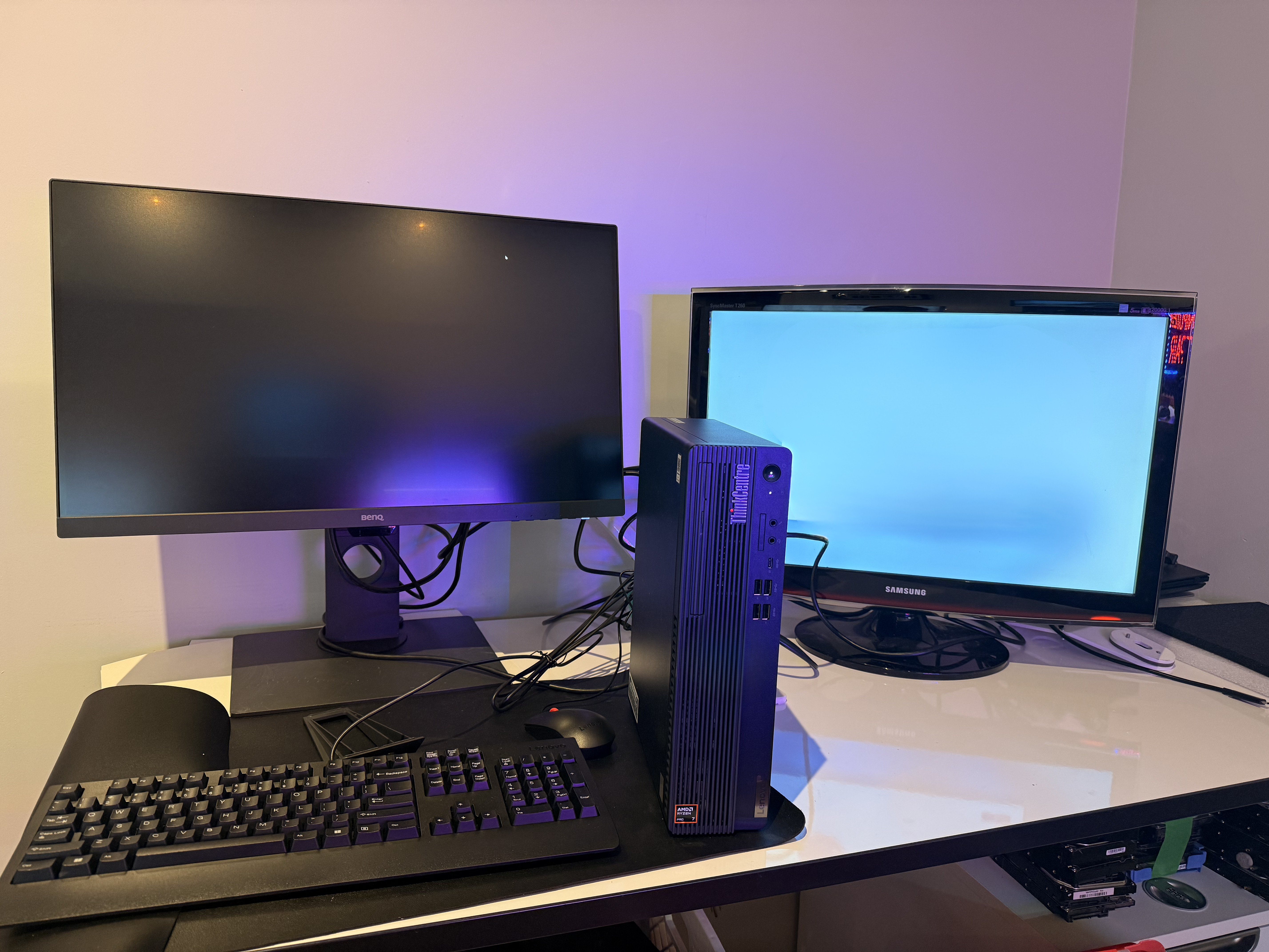 On the left, a BenQ monitor with a matte black screen is powered off, showing a faint purple reflection. On the right, an older, glossy Samsung SyncMaster T240 monitor is turned on, displaying a solid light blue screen. On the left, a BenQ monitor with a matte black screen is powered off, showing a faint purple reflection. On the right, an older, glossy Samsung SyncMaster T240 monitor is turned on, displaying a solid light blue screen.