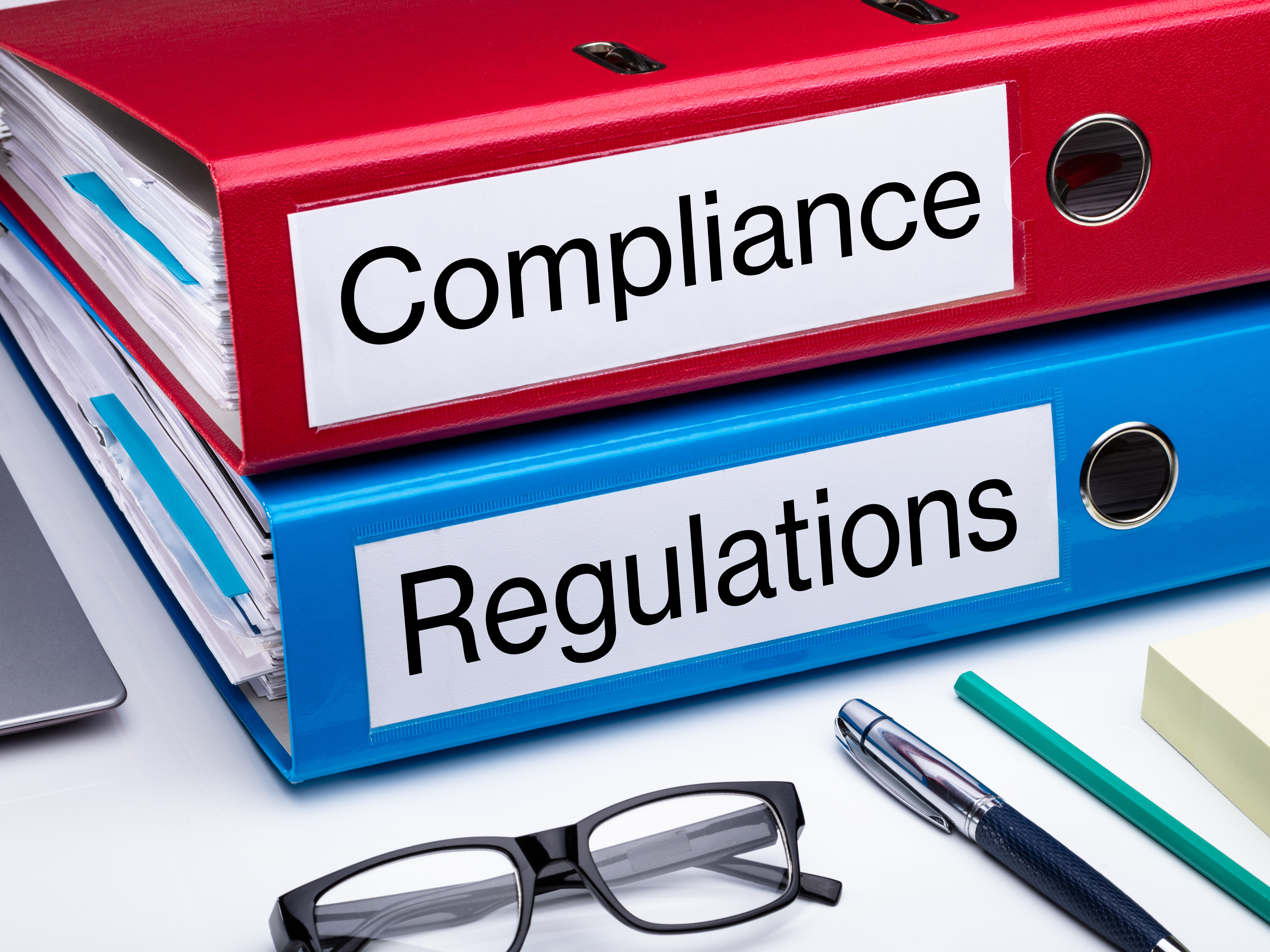 Close-up of red and blue office binders labeled "Compliance" and "Regulations" on a desk with documents, eyeglasses, and a pen, representing regulatory compliance, governance, policy management, and documentation for healthcare or business organizations.