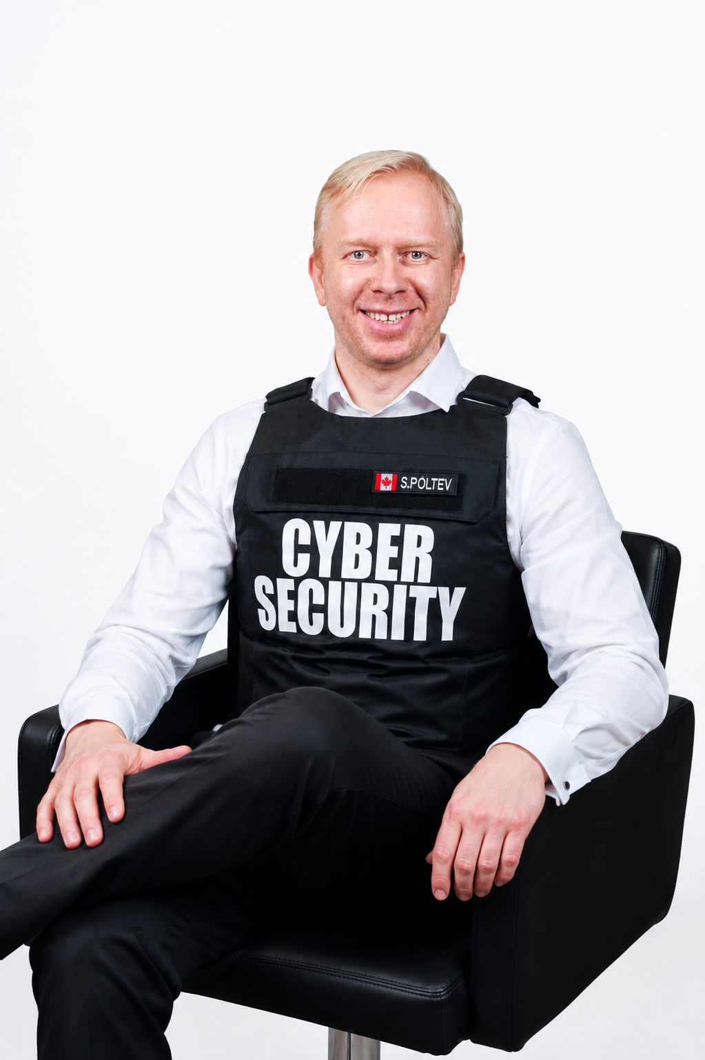 Professional portrait of a smiling cybersecurity expert seated in a modern black chair, wearing a white dress shirt and a black tactical vest labeled "CYBER SECURITY." The subject faces the camera with a confident, approachable expression against a clean white background, conveying trust, professionalism, and expertise in cybersecurity.