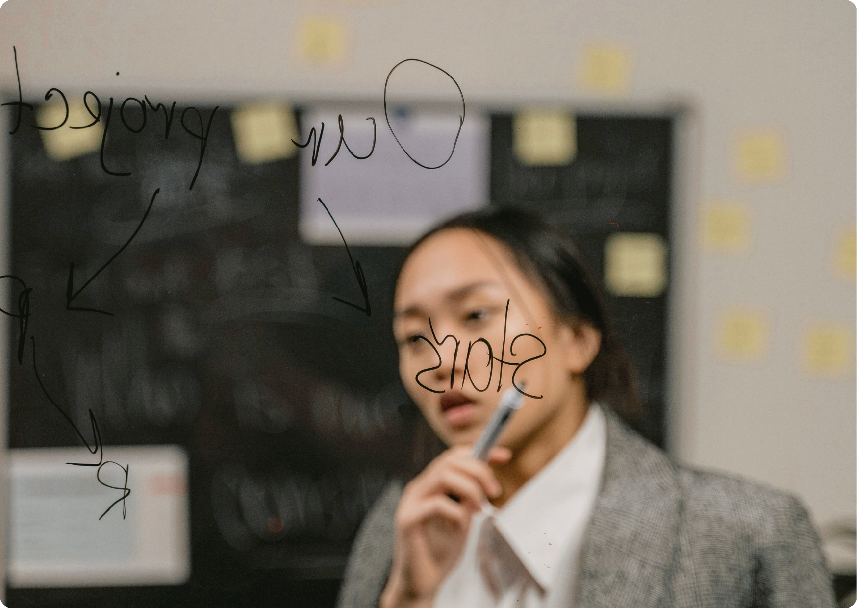 Blog Aude Compose - Femme asiatique en pleine réflexion sur un sujet, un crayon sur les lèvres, des écritures sur un tableau translucide, des post-it, un tableau à la craies au fond