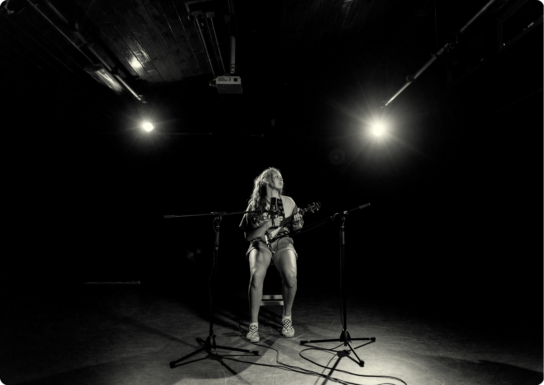 Blog Aude Compose - Femme seule aux longues dreadlocks blondes, avec une mini bass, assise sur un tabouret, des micros de studios, des projecteurs à faible luminosité et la pénombre