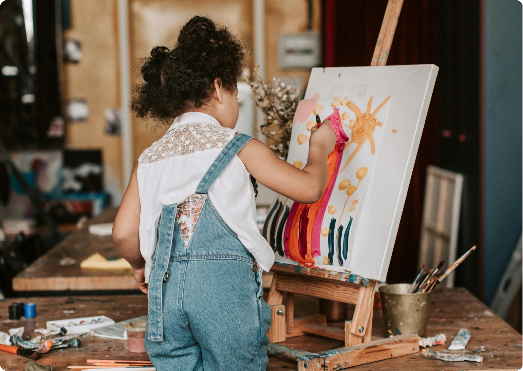 Blog Aude Compose - Enfant frisée de dos debout sur une chaise fait de la peinture, un chevalet posé sur une table