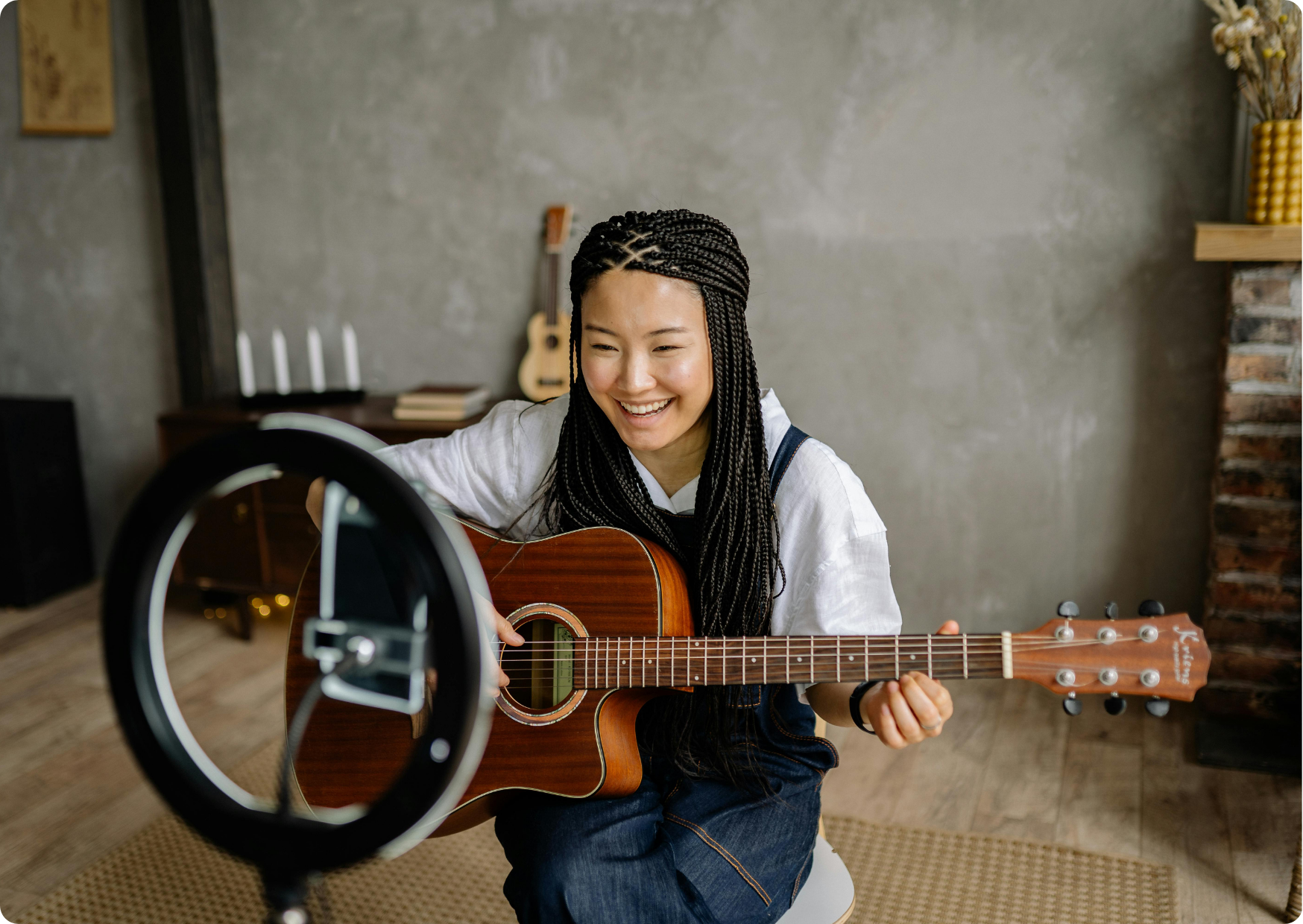 Blog Aude Compose - Femme asiatique tressée, sourit face à son smartphone entouré d'un ring, avec une guitare, dans un grande pièce, un ukulélé au fond.png