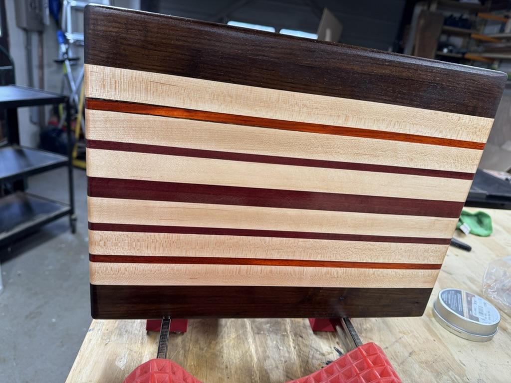 Handcrafted wooden cutting board with alternating light and dark wood stripes, including thin red accent strips, clamped on a workbench in a workshop.