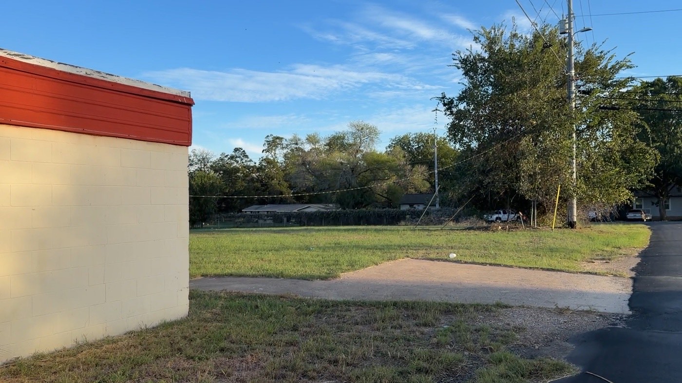 Side of the existing building with open, level grass and a paved drive beside it