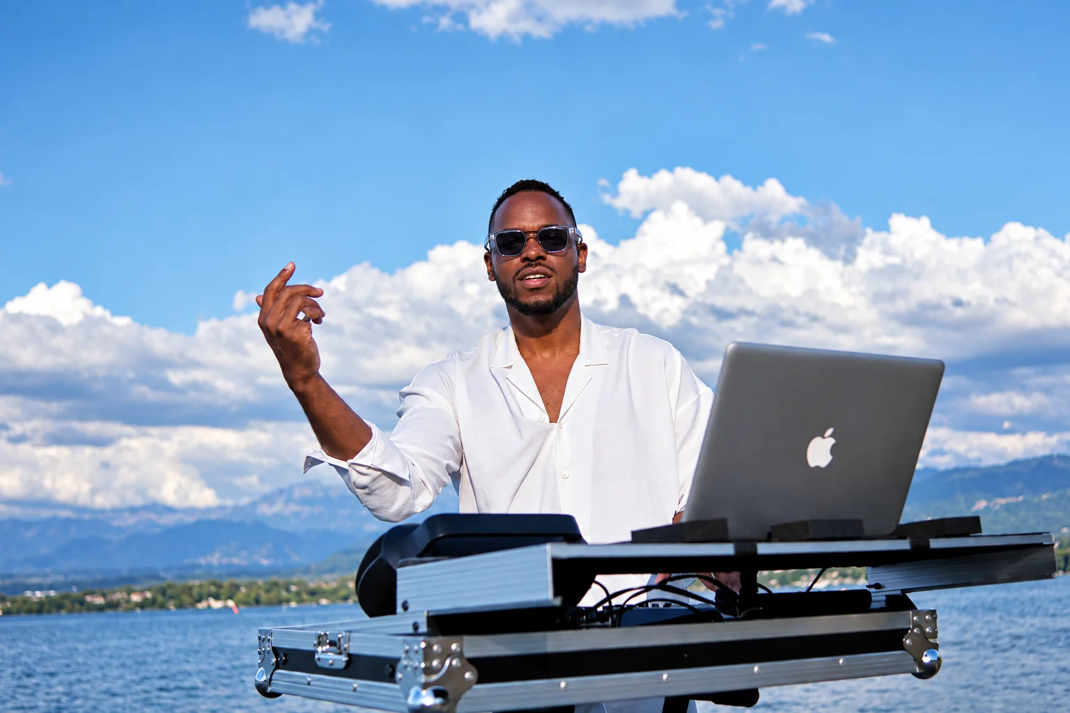 Un homme portant des lunettes de soleil et une chemise blanche se tient devant du matériel de DJ avec un MacBook, faisant des gestes près d’un lac.