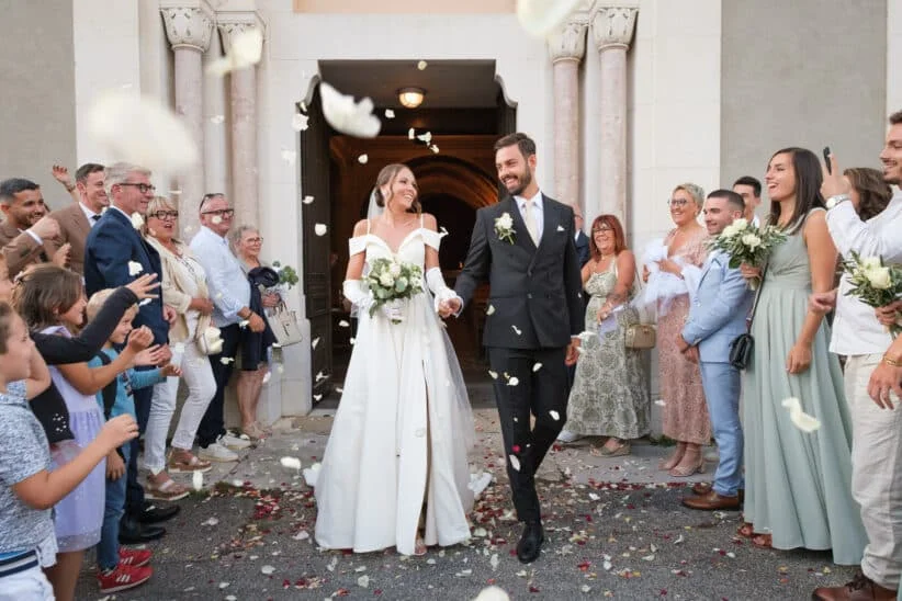 Un couple de jeunes mariés souriant sort d’un bâtiment en se tenant la main, tandis que les invités au mariage lancent des pétales blancs autour d’eux.