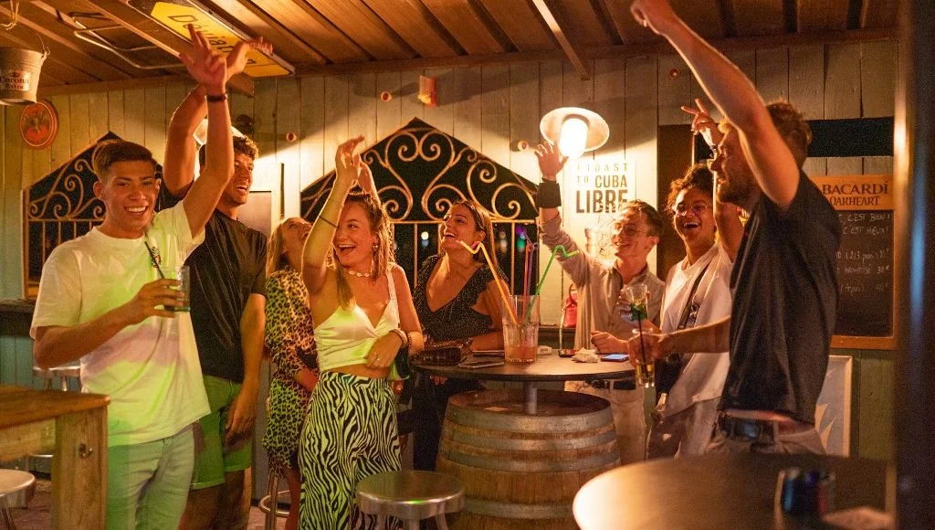 Un groupe d’amis joyeux, les mains levées, célèbre et rit dans un bar faiblement éclairé, avec des boissons posées sur une table en tonneau.