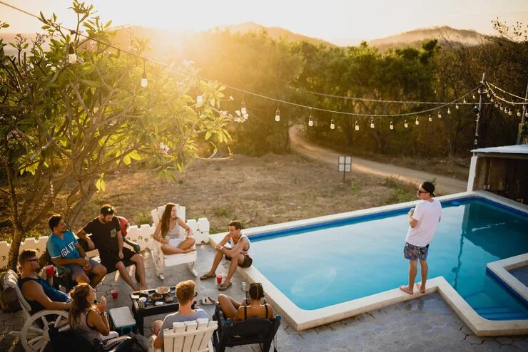 Un groupe d’amis se détendant au bord d’une piscine en plein air à l’heure dorée, avec des guirlandes lumineuses et des collines verdoyantes en arrière-plan.