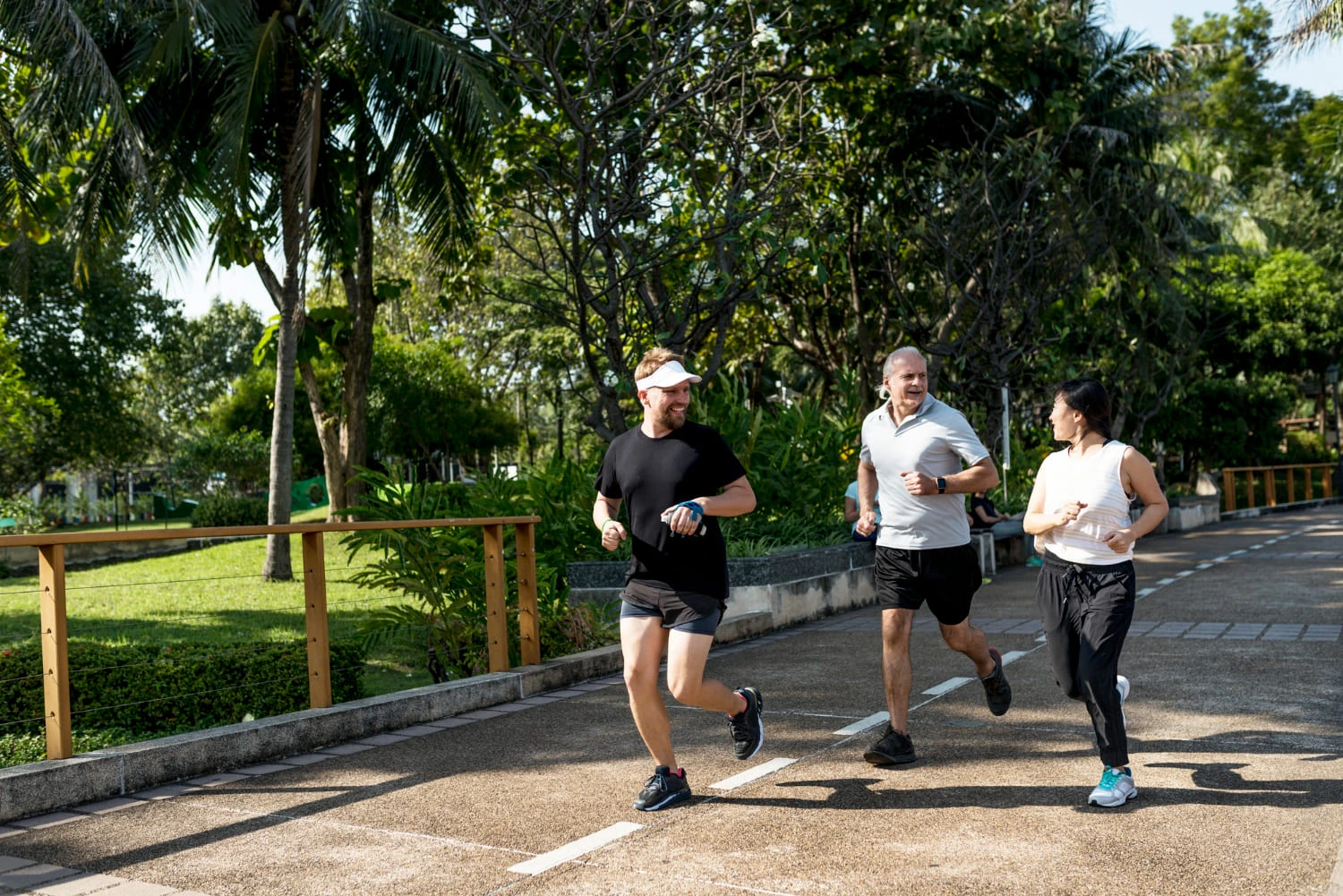 Tres adultos corriendo al aire libre en un parque soleado, haciendo ejercicio juntos en un sendero pavimentado.