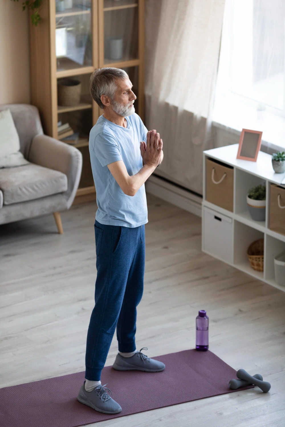 Hombre mayor de pie sobre una esterilla en casa, realizando una postura de meditación o respiración con las manos juntas frente al pecho.