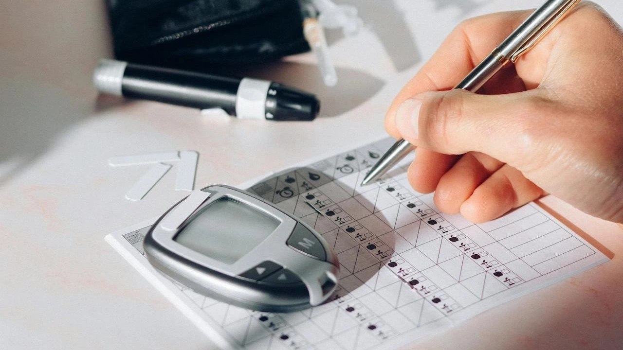 Persona registrando niveles de glucosa en una hoja mientras un glucómetro y un estuche con suministros para diabéticos descansan sobre la mesa.