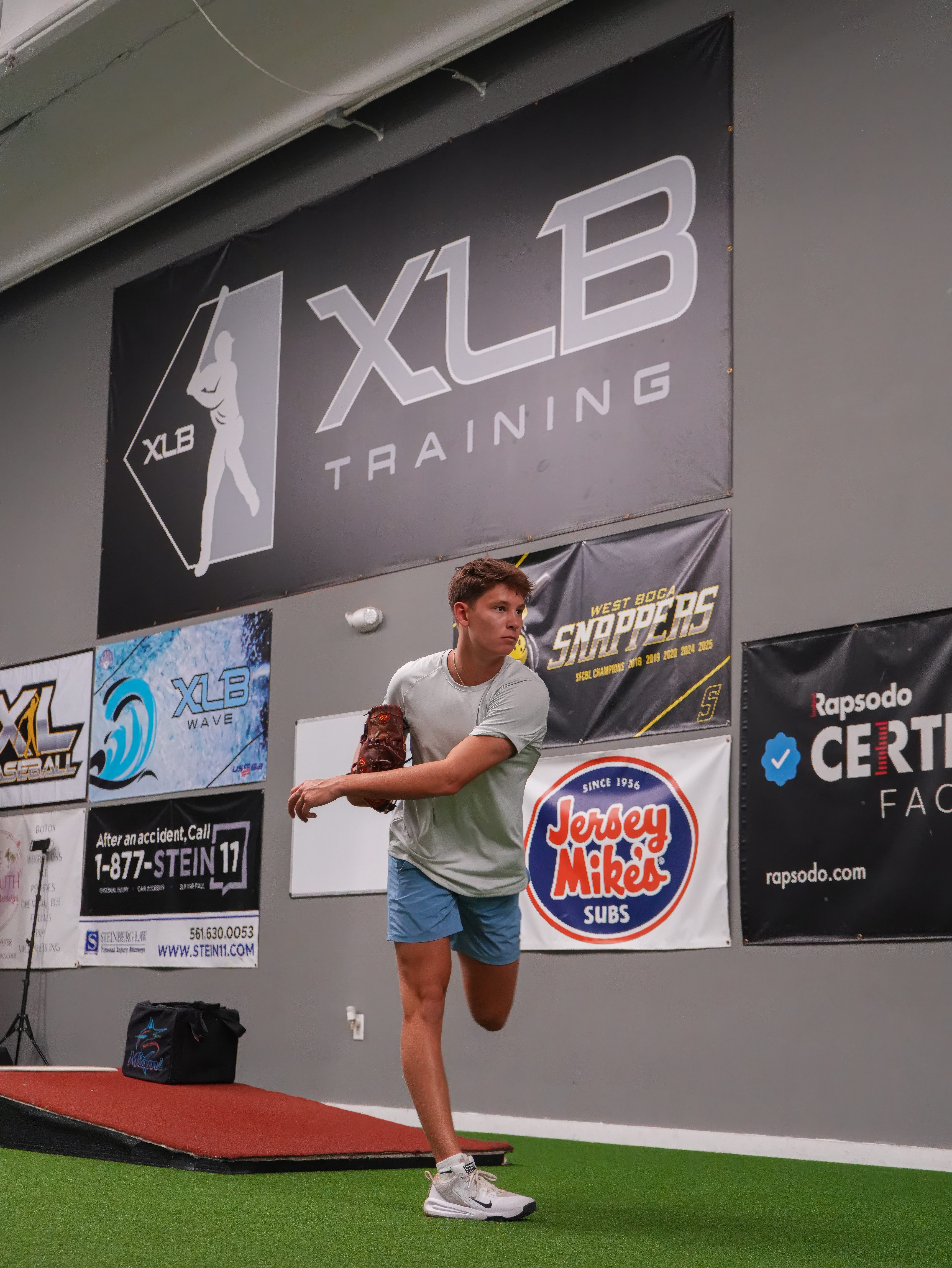 Young pitcher finishing his delivery in front of the XLB Training banner at the Rapsodo certified facility