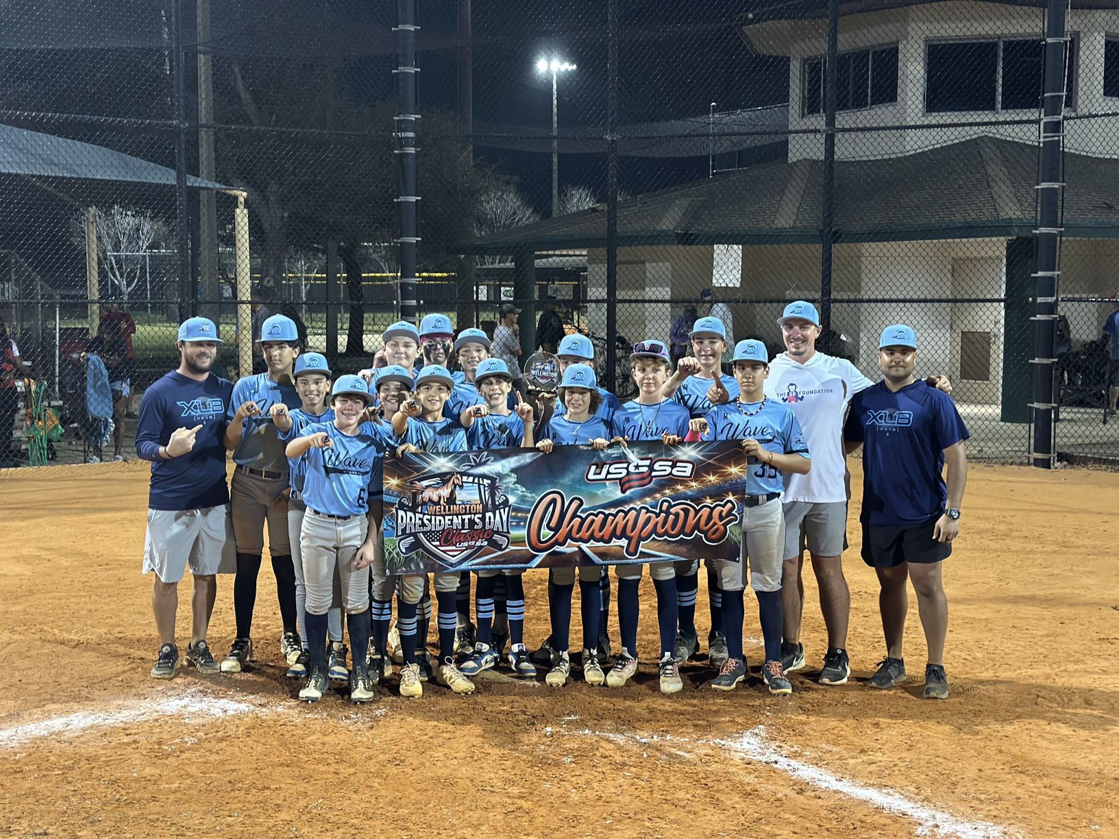 An XLB Wave youth travel baseball team and its coaches holding a tournament champions banner in Palm Beach County, Florida
