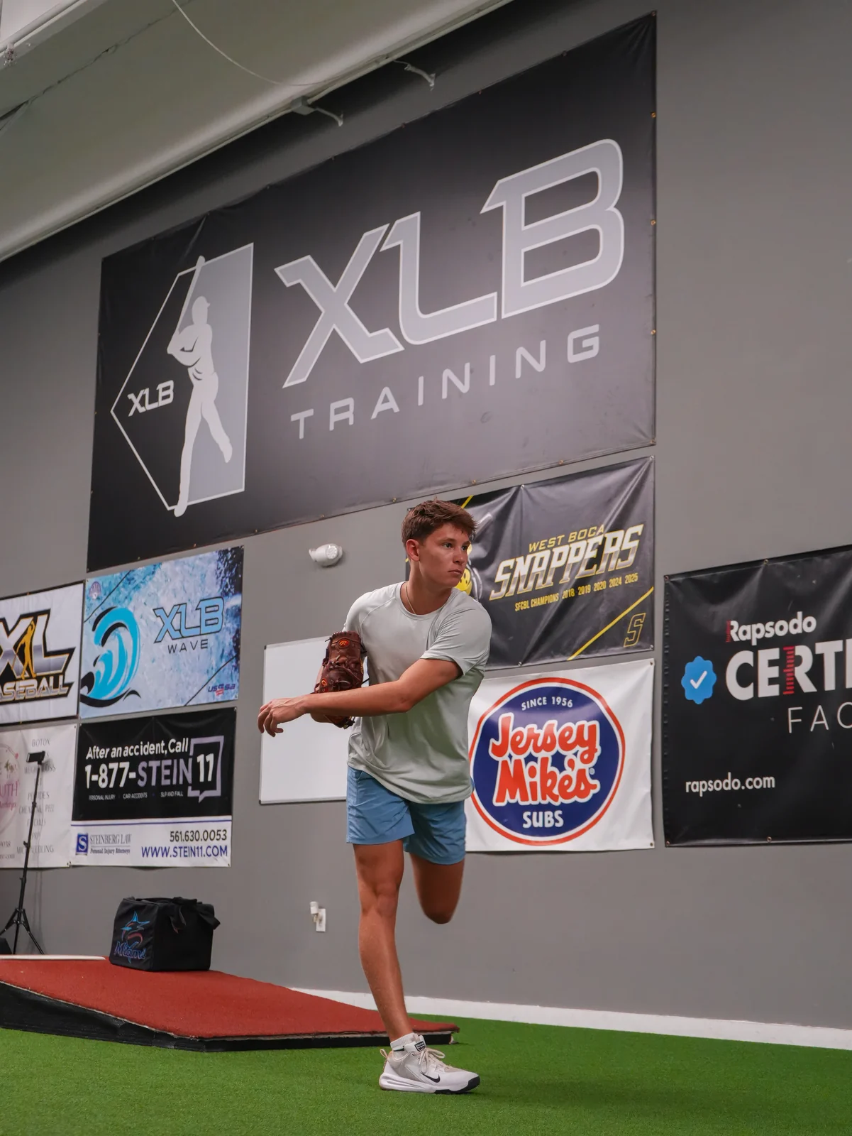 Young pitcher finishing his delivery in front of the XLB Training banner at the Rapsodo certified facility