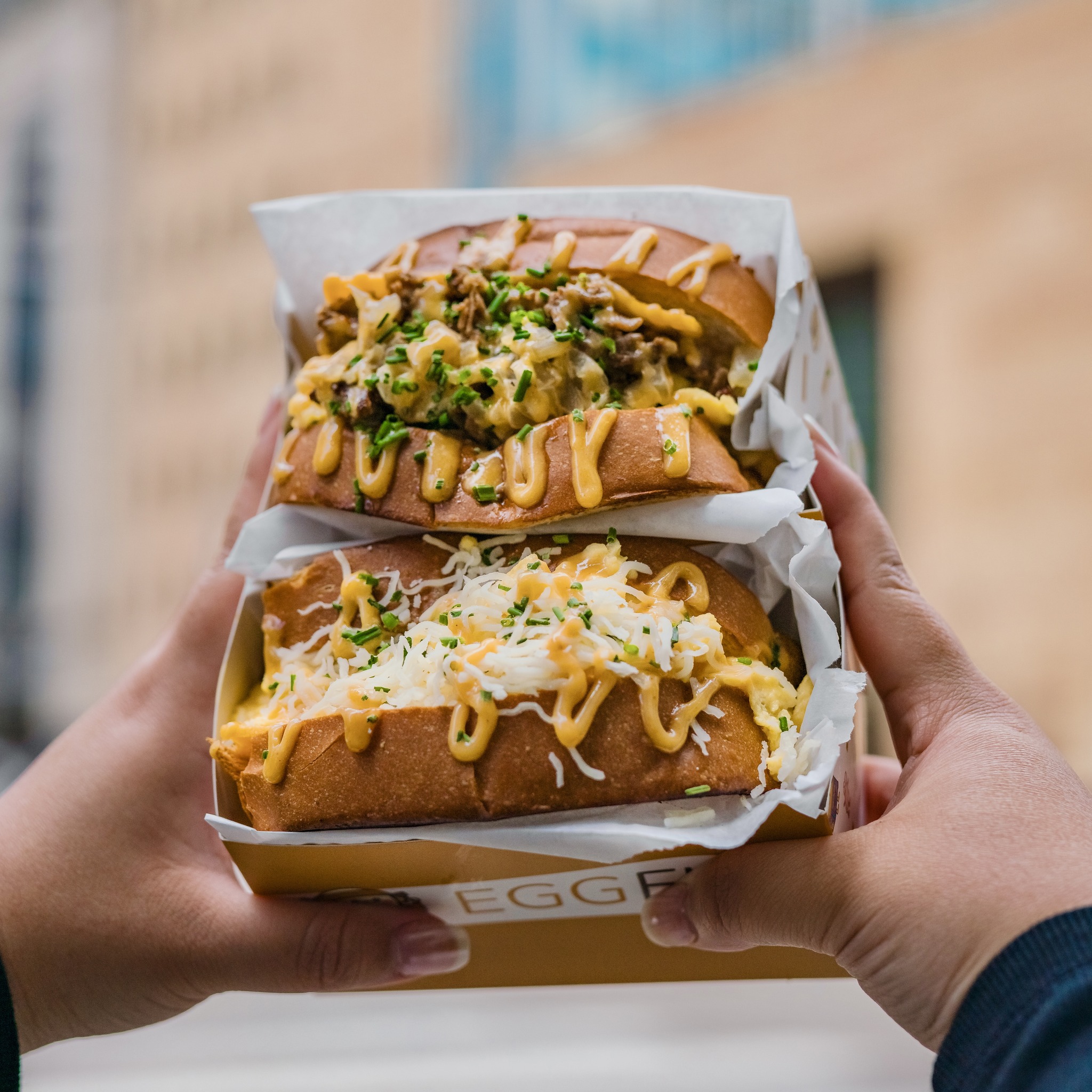Hands holding two EGGFLIP egg sandwiches loaded with toppings