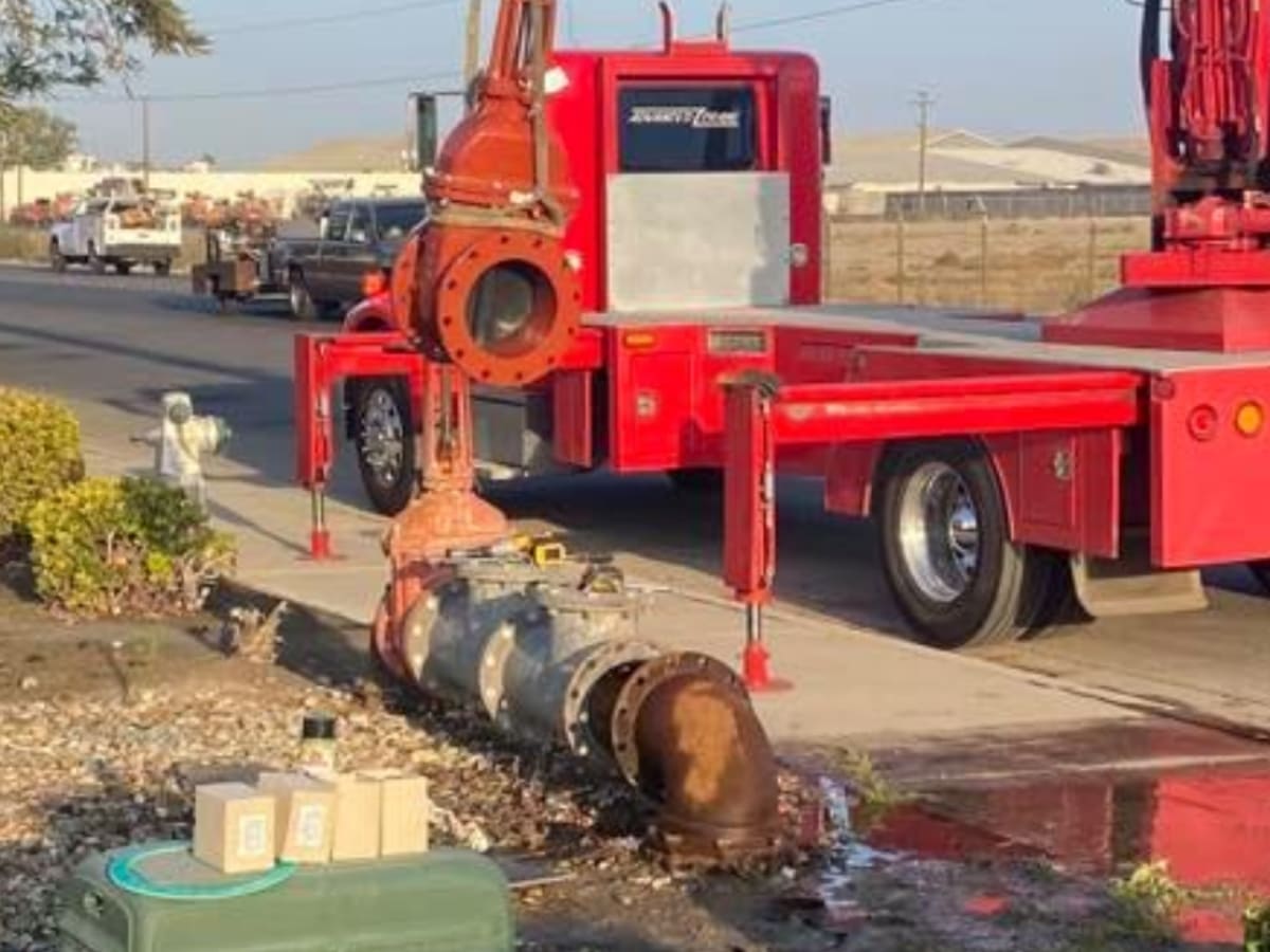 A large industrial valve system being removed with the help of a crane on a roadside, showcasing the heavy equipment involved in backflow repair.