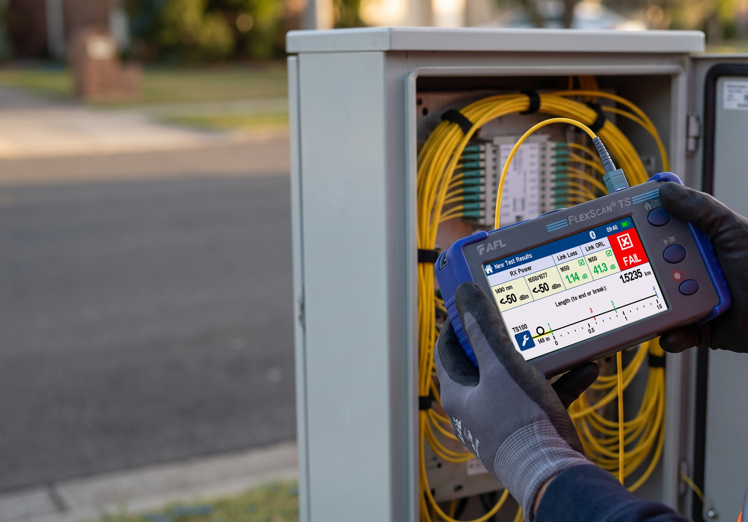 A field technician holding an AFL FlexScan fiber tester in front of an open fiber cabinet