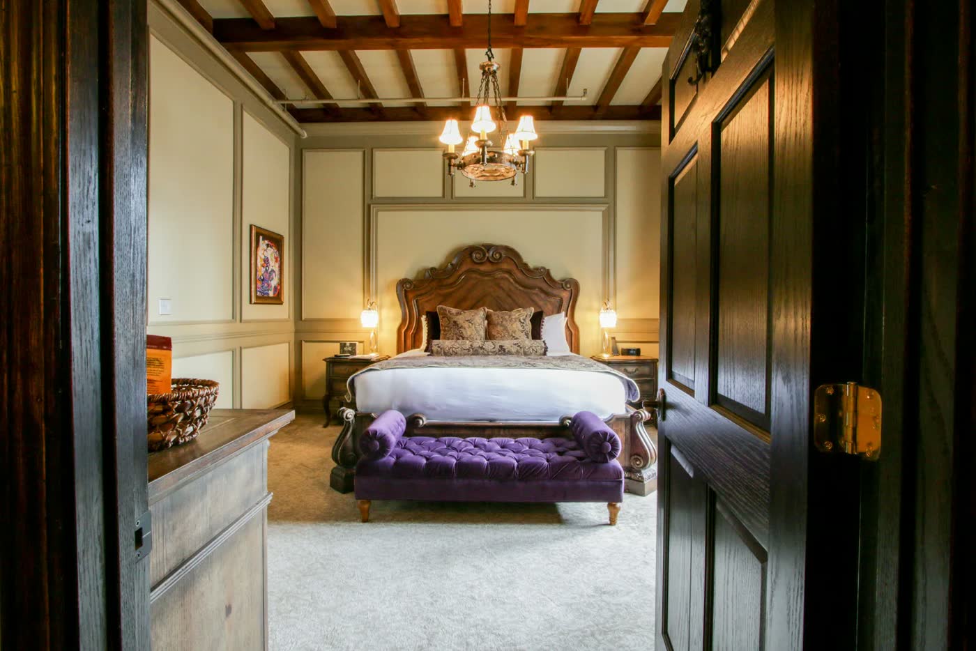 A suite seen through a carved doorway: chandelier, upholstered bed and tufted bench