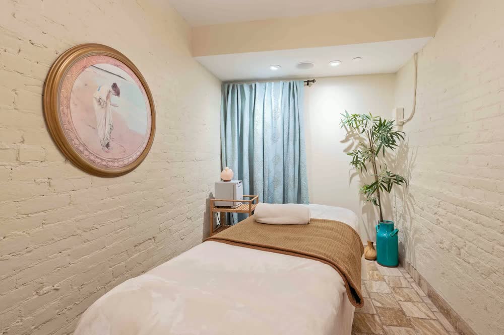 A treatment room with a made massage table, blue curtains and a potted palm