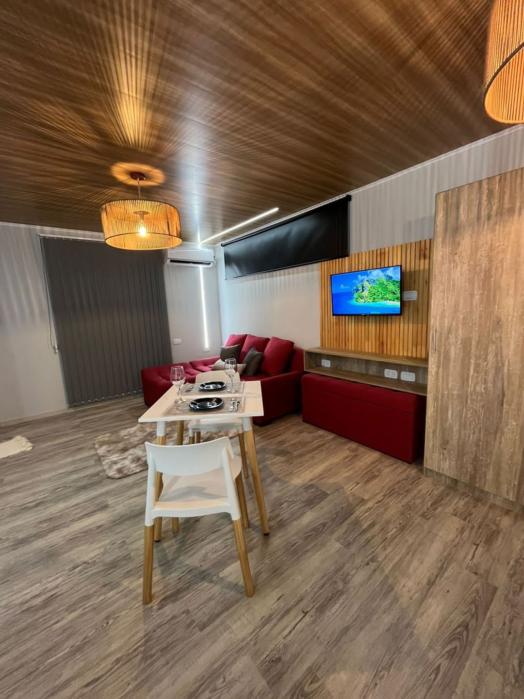 Living con sillón de terciopelo rojo, TV, lámparas de ratán y mesa para dos